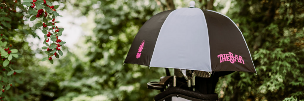 The top of a golf bag with clubs in it and a black and white golf bag umbrella with pink lettering over the bag while it's sitting in a wooded area