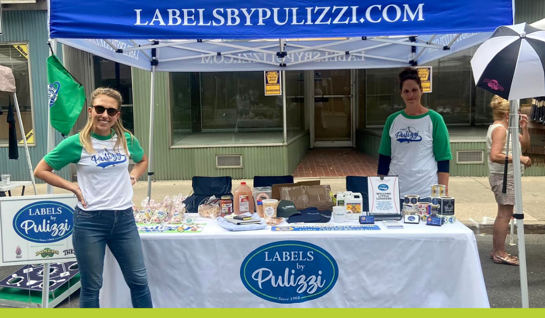 Labels by Pulizzi popup tent set up at an event with products on a table and two women dressed in a shirt with the brand logo and jeans smiling